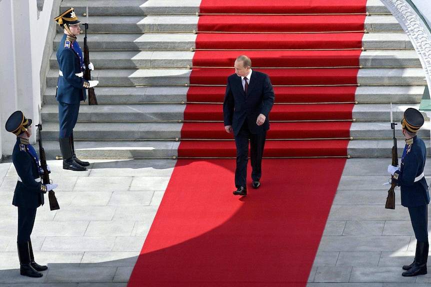 Russian president Vladimir Putin walking a red carpet while being watched by three guards.