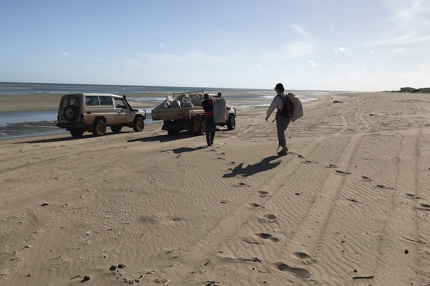 A photo of two volunteers trekking along a beach, towards two vehicles, carrying bags of waste.