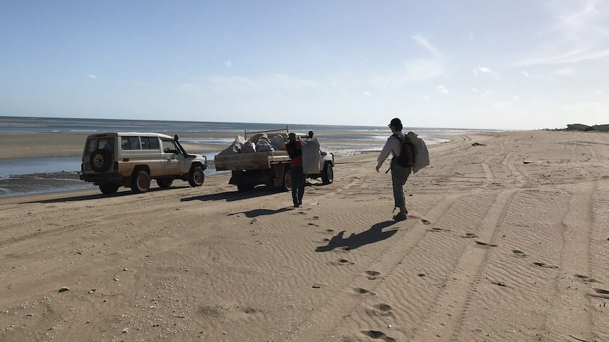 A photo of two volunteers trekking along a beach, towards two vehicles, carrying bags of waste.