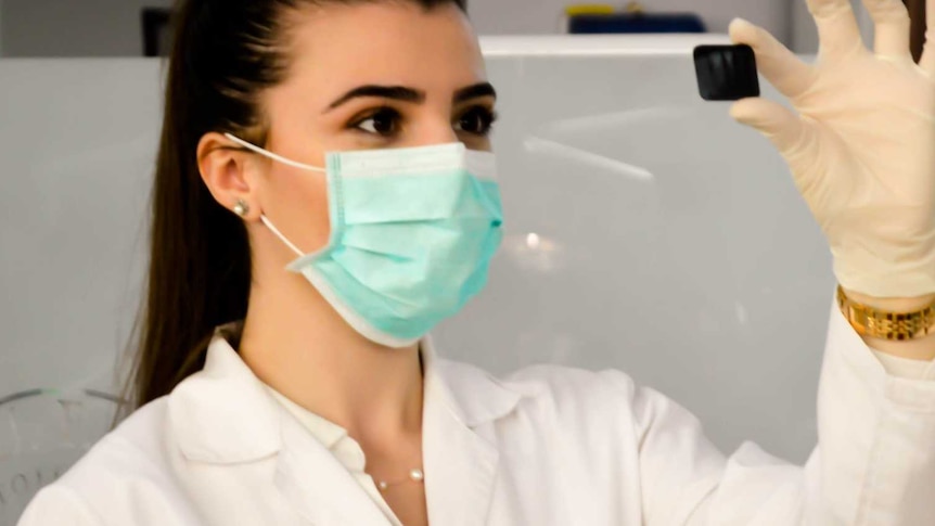 Woman in lab coat and mask in in medical setting examines small black slide