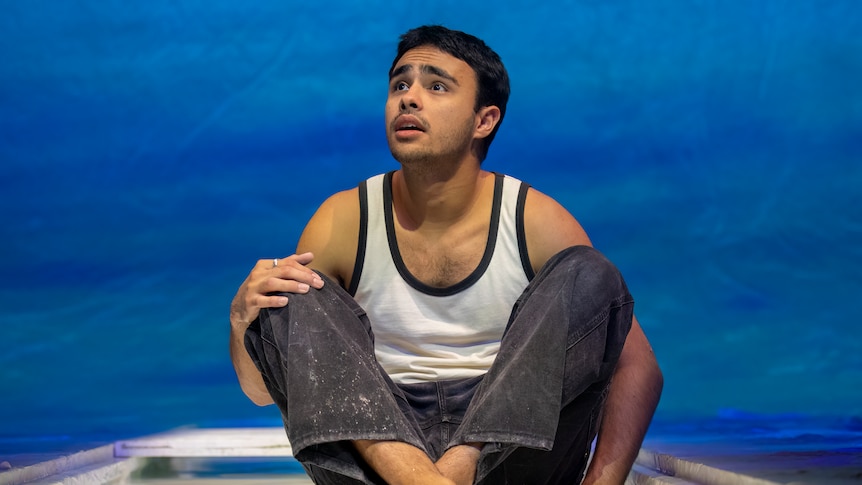 An Indigenous man, in his early 20s, wearing a singlet and jeans, sits cross-legged on stage. He looks upset, his chin raised.