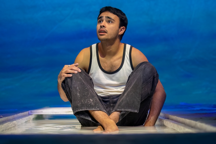 An Indigenous man, in his early 20s, wearing a singlet and jeans, sits cross-legged on stage. He looks upset, his chin raised.