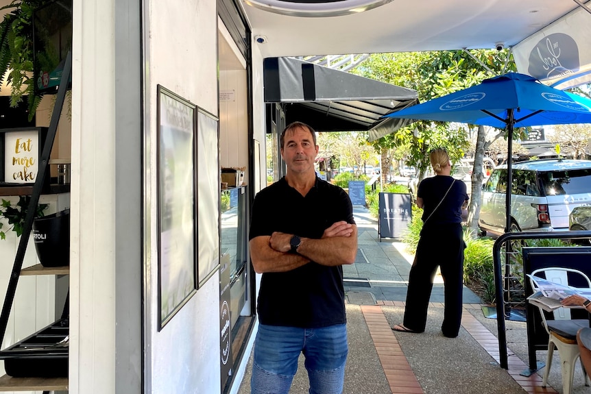 Man standing on footpath under a cafe sign with arms crossed. 