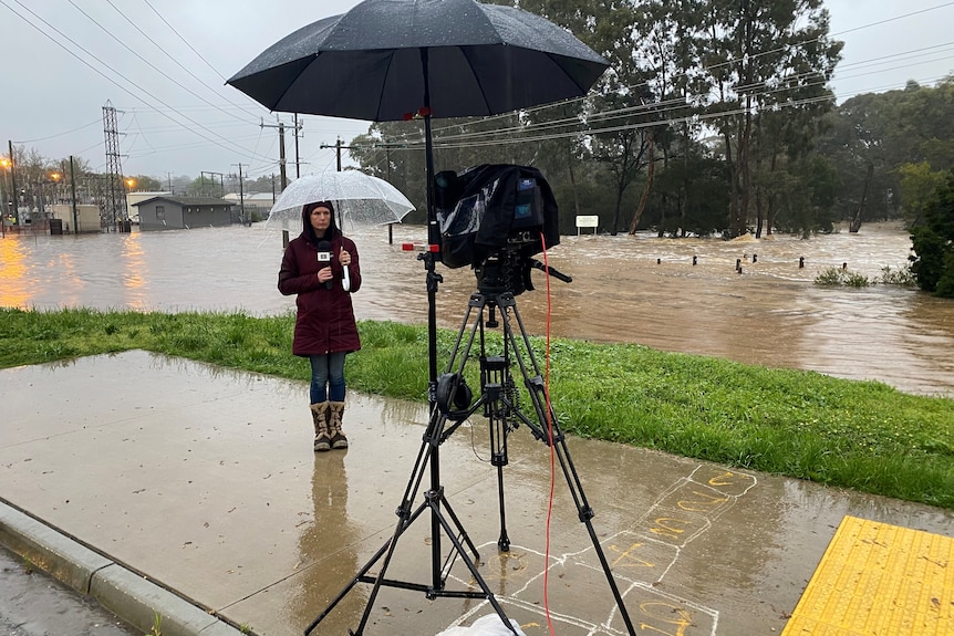 Woman standing under and umbrella, holding a microphone as camera on a tripod films her and floods in background.