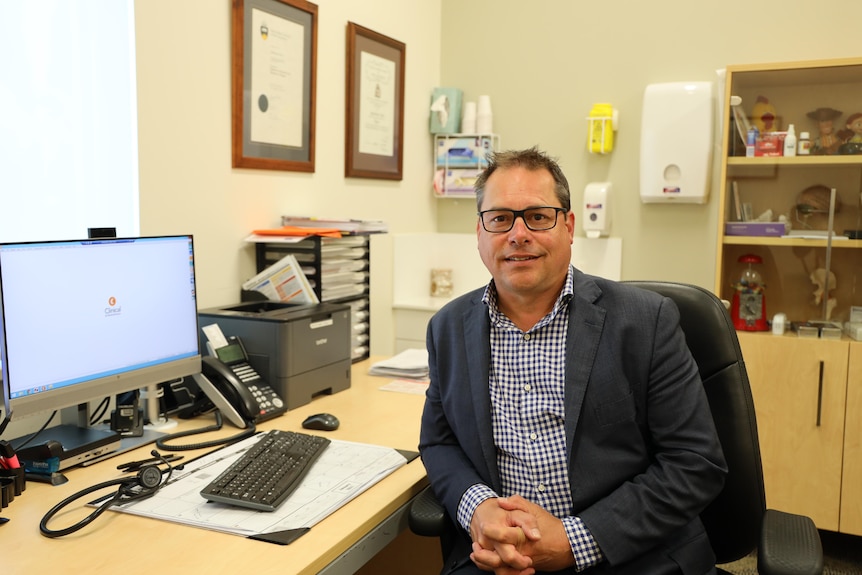 A man in a casual suit jacket and glasses sits at a computer desk, medical equipment behind him.