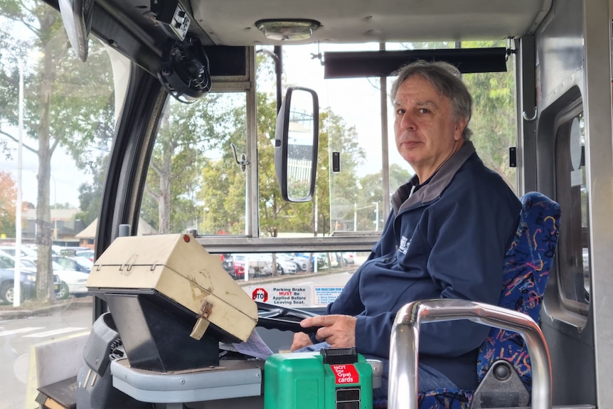 Bob Gioia sits in the driver's seat wearing a blue jumper, driving an old bus and looking at the camera.