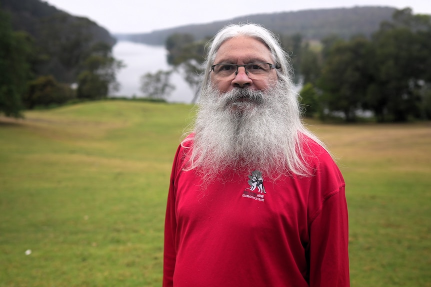 Uncle Gerry has a long white beard and glasses and is wearing a bright red shirt. The shoalhaven river is behind him
