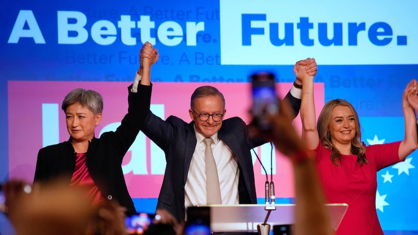 Wide shot of three people linking hands on a stage.