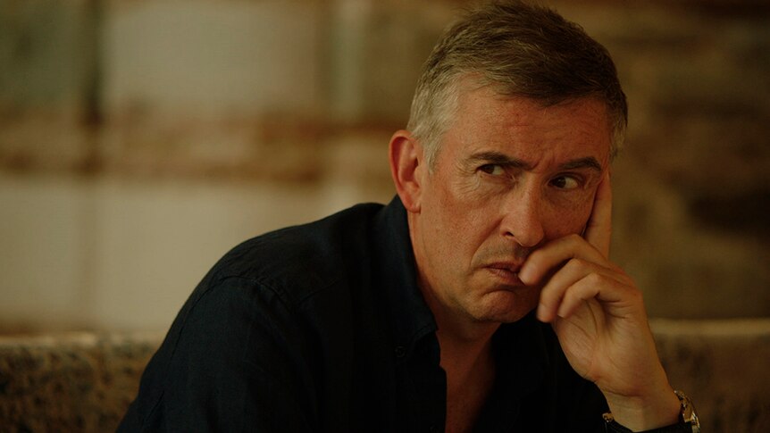 A man in black shirts with short greying dark hair makes serious expression and rests chin in hand, background is out of focus.