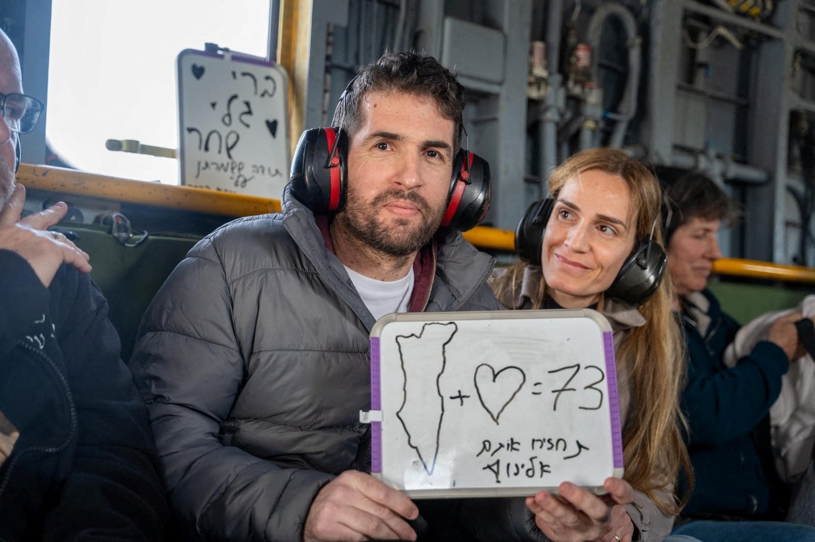 A smiling man and woman, wearing earmuffs, sit on a plane. The man holds a sign. Words in Hebrew read, "Bring them back to us"