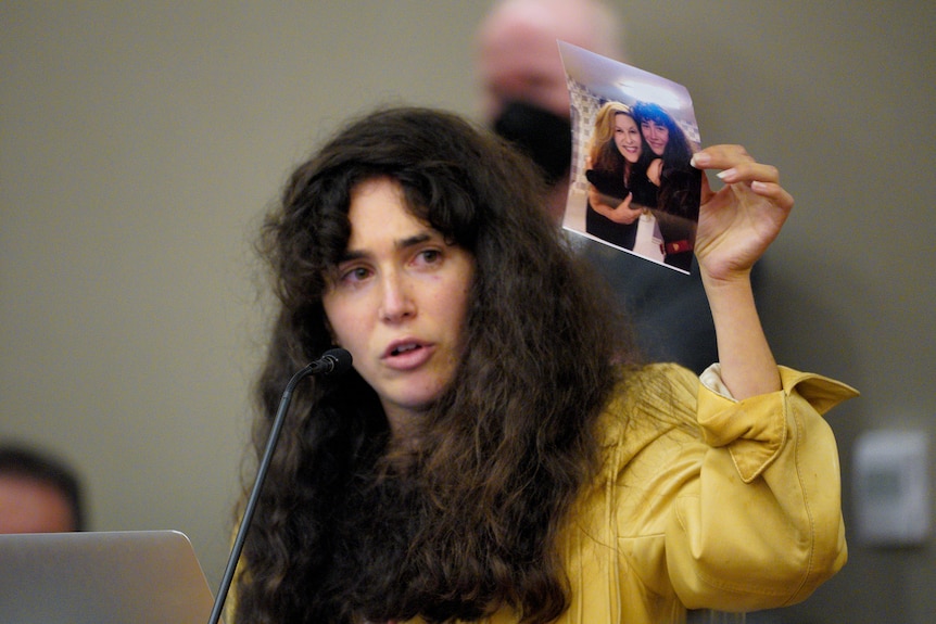 a woman with long brown hair wearing a yellow leather jacket holds up a photo of herself and an older woman.