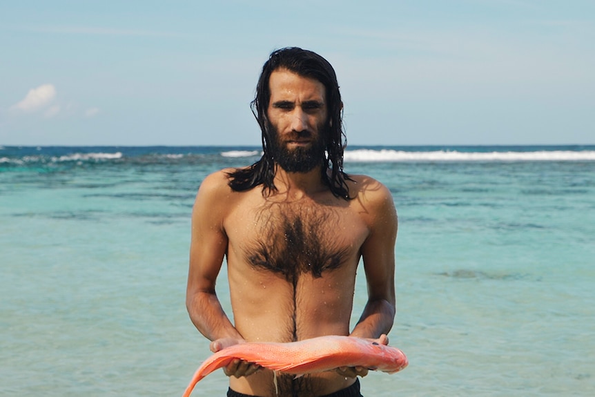 Colour still of Behrouz Boochani holding a fish by the ocean from video artwork Remain by artist Hoda Afshar.