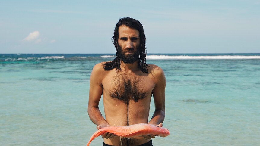 Colour still of Behrouz Boochani holding a fish by the ocean from video artwork Remain by artist Hoda Afshar.