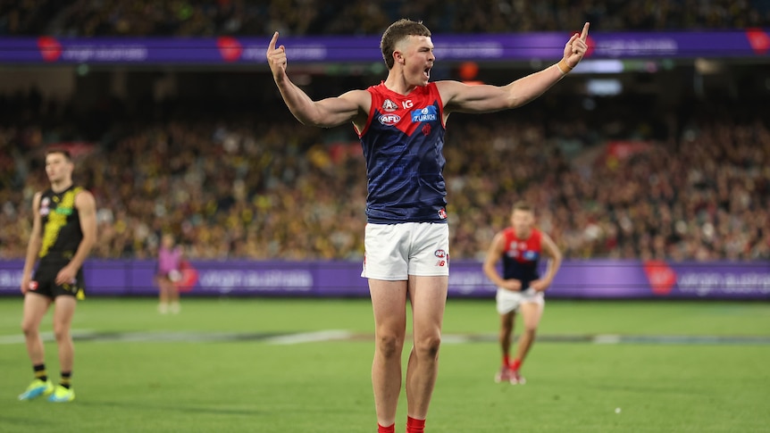 A Melbourne AFL player stands on tiptoe and roars in celebration pointing his fingers in the air.