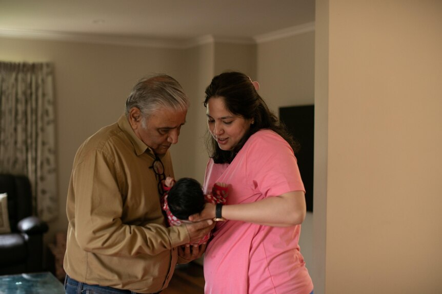 An older man with greying hair takes a baby from a young woman wearing pink tee. Both look at the baby.