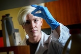 Alea Rose wears a lab coat and blue gloves, as she holds a pipette in front of her eyes