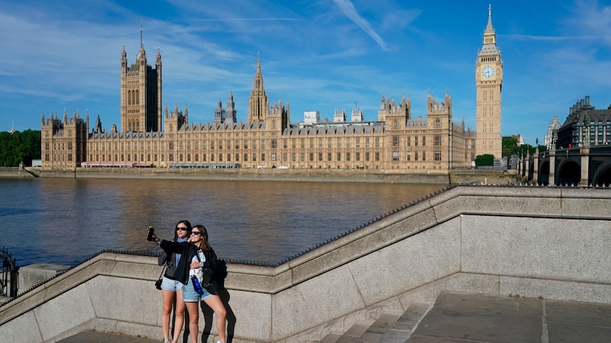 Two women in sunglasses take a selfie with Big Ben in the background, beside the Thames river.