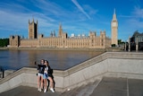 Two women in sunglasses take a selfie with Big Ben in the background, beside the Thames river.