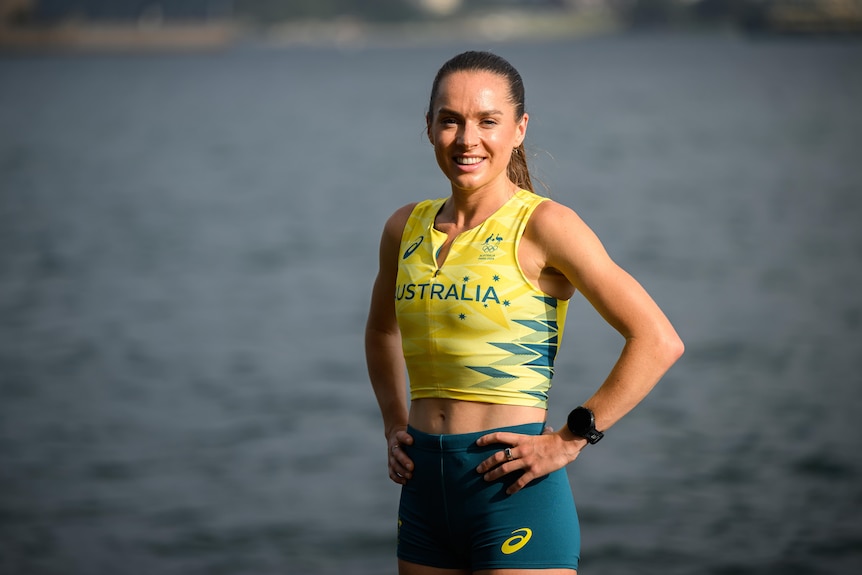 A female runner, Izzi Batt-Doyle, is wearing an Australian Olympic team uniform, posing with hands on her hips, smiling.