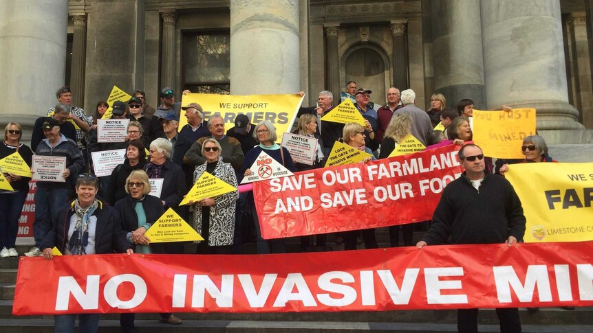 Men and women on the stone steps of Parliament House hold red signs with white writing saying No Invasive Mining
