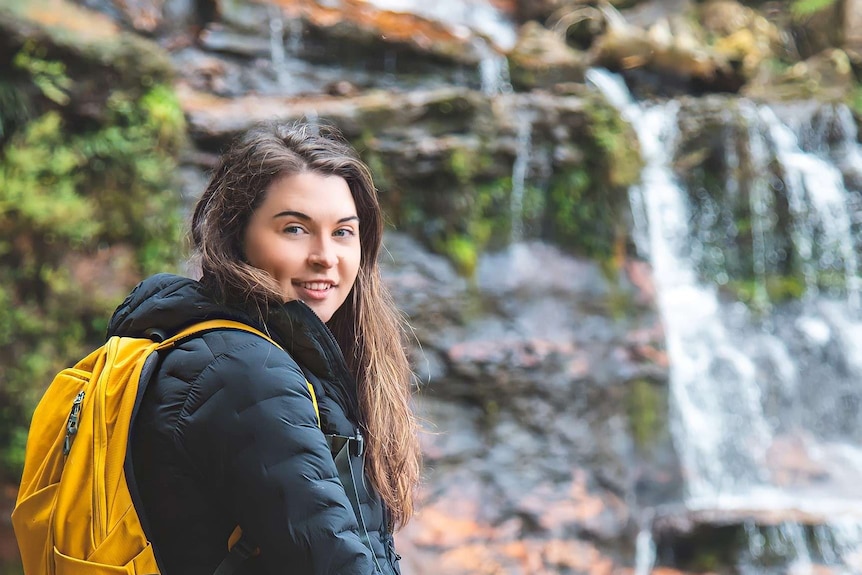 A young woman with long brown hair smiles at the camera next to a waterfall