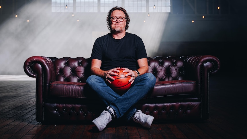 Luc sits on a purple couch wearing a black t-shirt and jeans and holding a basketball.