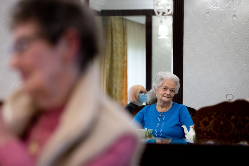 An older woman sits at a table drinking tea and chatting with a group of other elderly women.