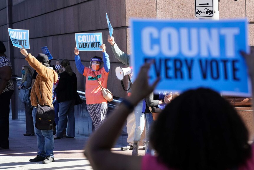 A number of people wearing face shields and masks hold up signs saying Count Every Vote outside a building.