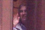 A man in a grey jacket smiles and waves goodbye to a woman from a partially open door.