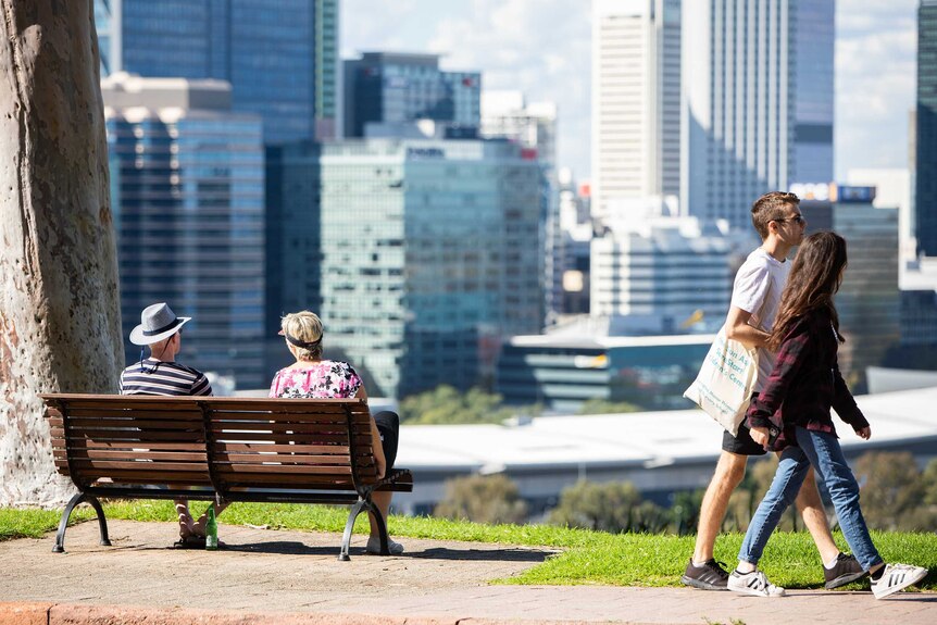 An older couple sit on a bench in Kings Park overlooking the Perth city as a younger couple walk past.