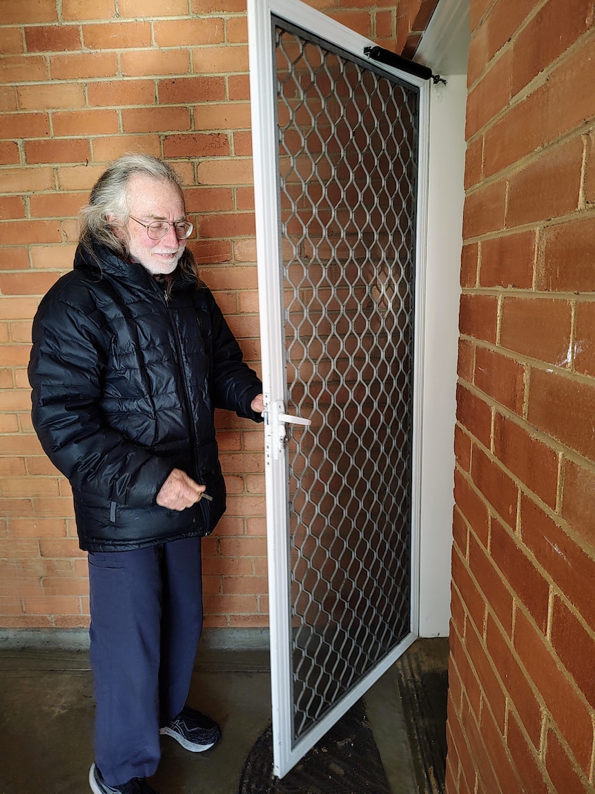 A man with grey hair and black jacket holds a key to a closed door near an open screen door.