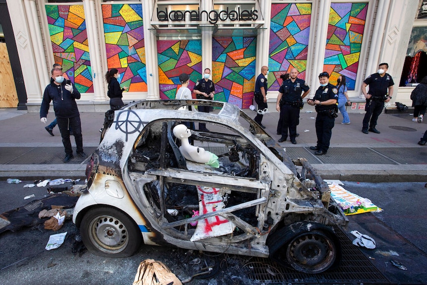 You view a burnt-out Smart car with a row of New York police officers looking on, next to a bright shop window display.