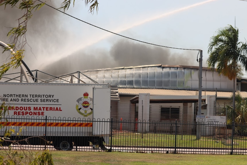 Se rocía agua en el techo de un edificio de ladrillo blanco.  El humo se eleva desde arriba. 