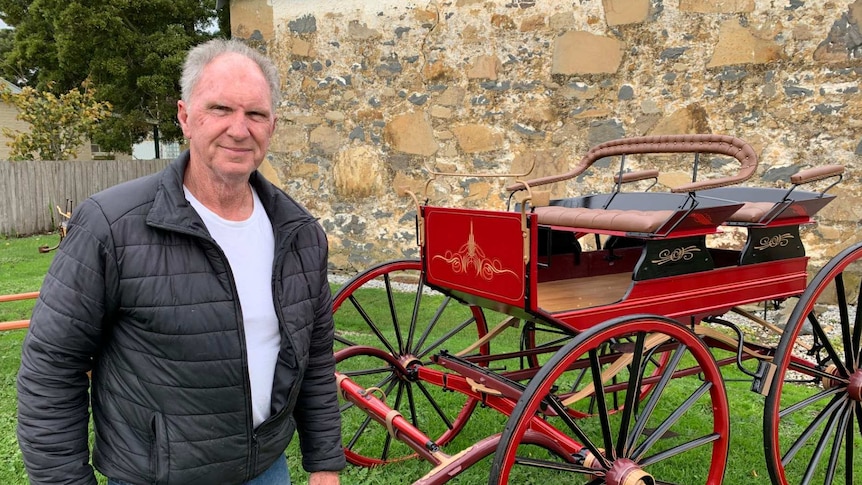 Rick Anderson poses for a photo in front of a red horse-drawn carriage he has restored.