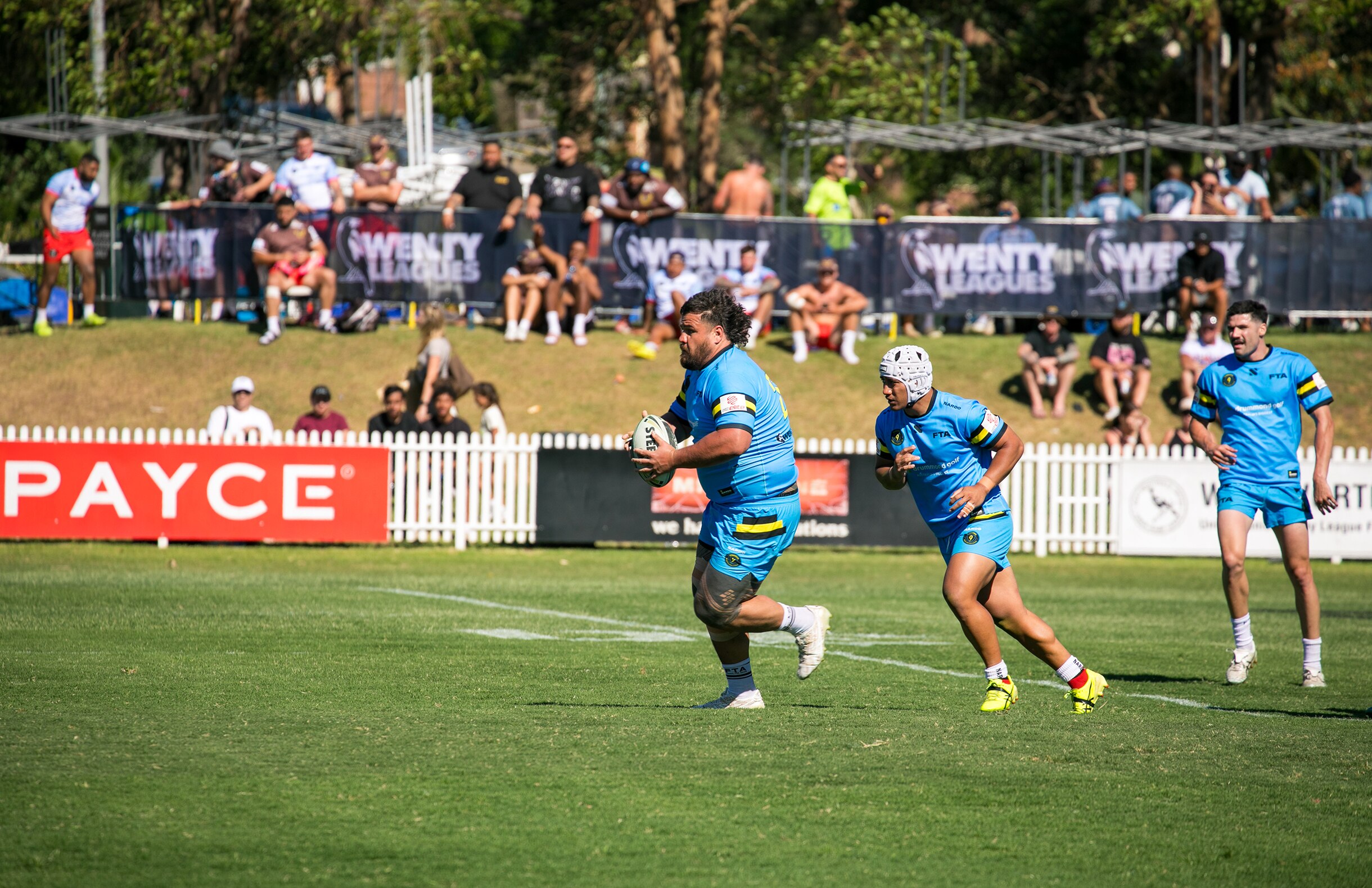Man in aqua matching football set runs on the rugby league field holding a ball.  He has a traditional Samoan tattoo on his legs