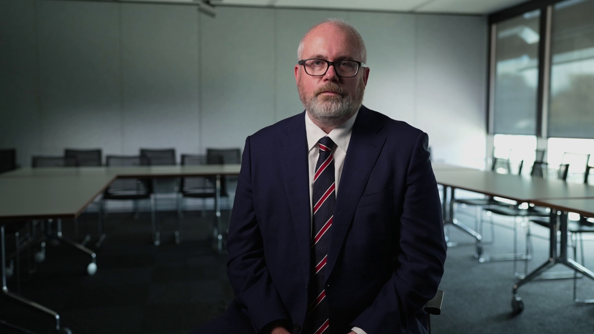 A balding man wearing a navy suit, white shirt, red and navy tie and glasses sits in a nondescript meeting room.