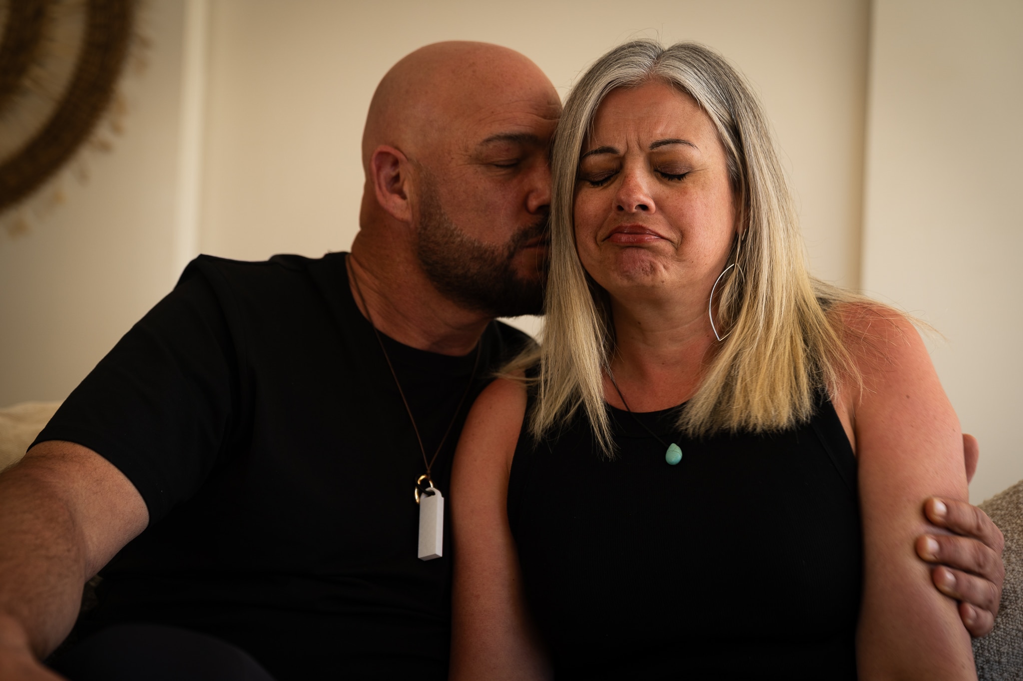 A man and a woman sit on a counch he has a beard and bald head and is kissing her cheek, she is crying.