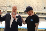 Michael Gudinski talks into a microphone while Ed Sheeran stands next to him smiling.