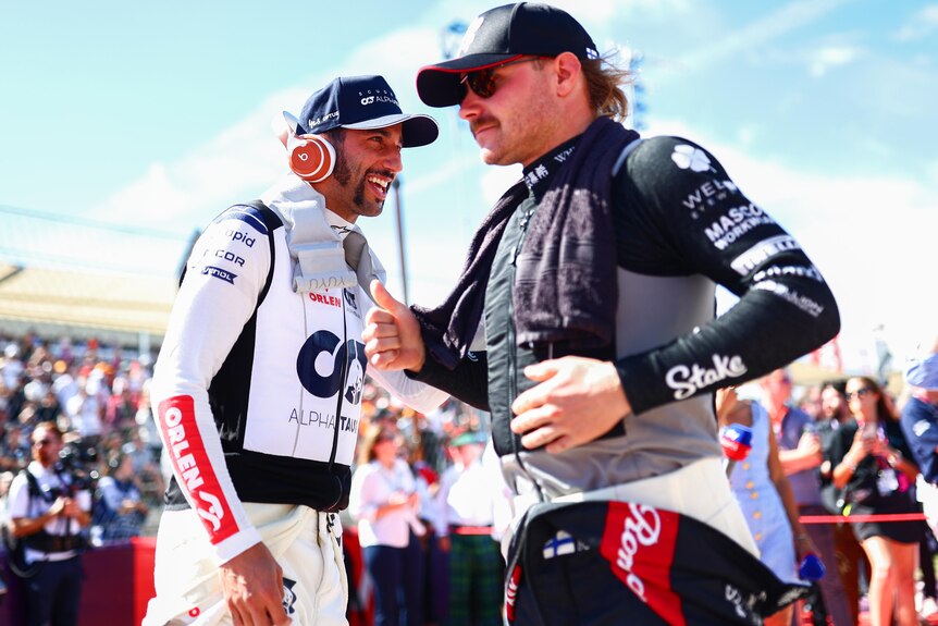 Two F1 drivers in their race suits, not helmets, both wearing caps, talking on the grid