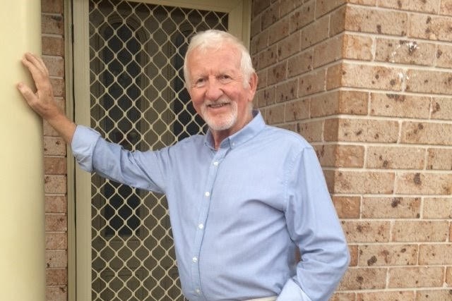 An older man wearing a blue shirt stands outside a doorway.