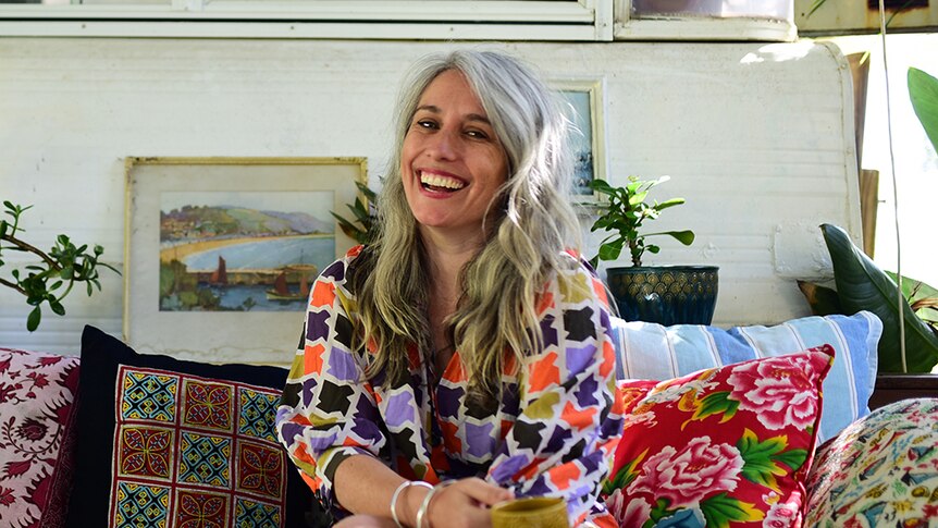 A woman with long, silver hair sitting on a couch with colourful cushions.