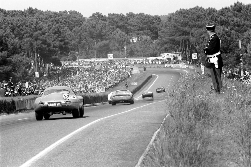 A police officer on the right of screen watches cars go down the back straight at Le Mans in a black and white photo.