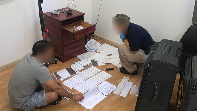 Two people with blurred faces are sitting on the floor in a house looking over documents.