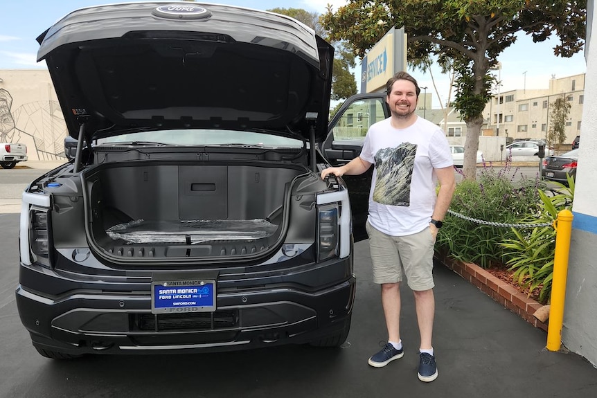 A man stands next to a big vehicle with it's bonnet open showing a battery.