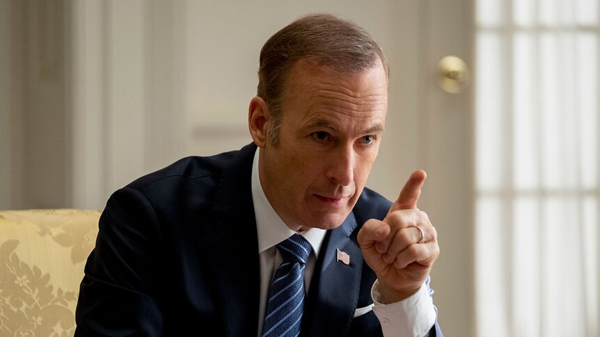 Colour close-up still of Bob Odenkirk wearing suit, seated and pointing finger in naturally lit room in 2019 film Long Shot.