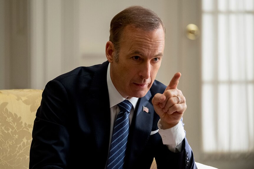 Colour close-up still of Bob Odenkirk wearing suit, seated and pointing finger in naturally lit room in 2019 film Long Shot.