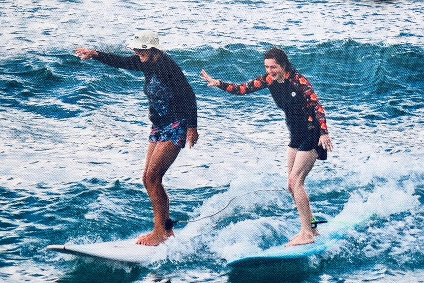 Two female surfers, one old, the other one younger, are standing on their boards laughing as they catch a wave together.