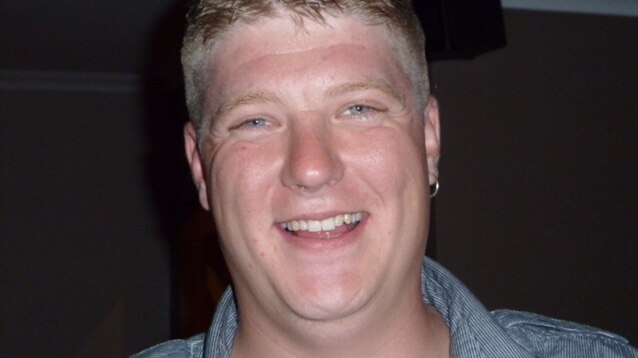 Anthony, wearing a blue-stripped collared shirt and an earing in one ear, smiles at the camera.