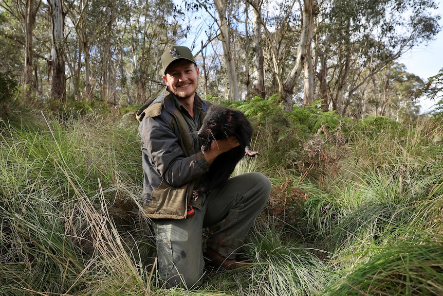 Man in dark green jacket and hat, smiles with Tasmanian devil