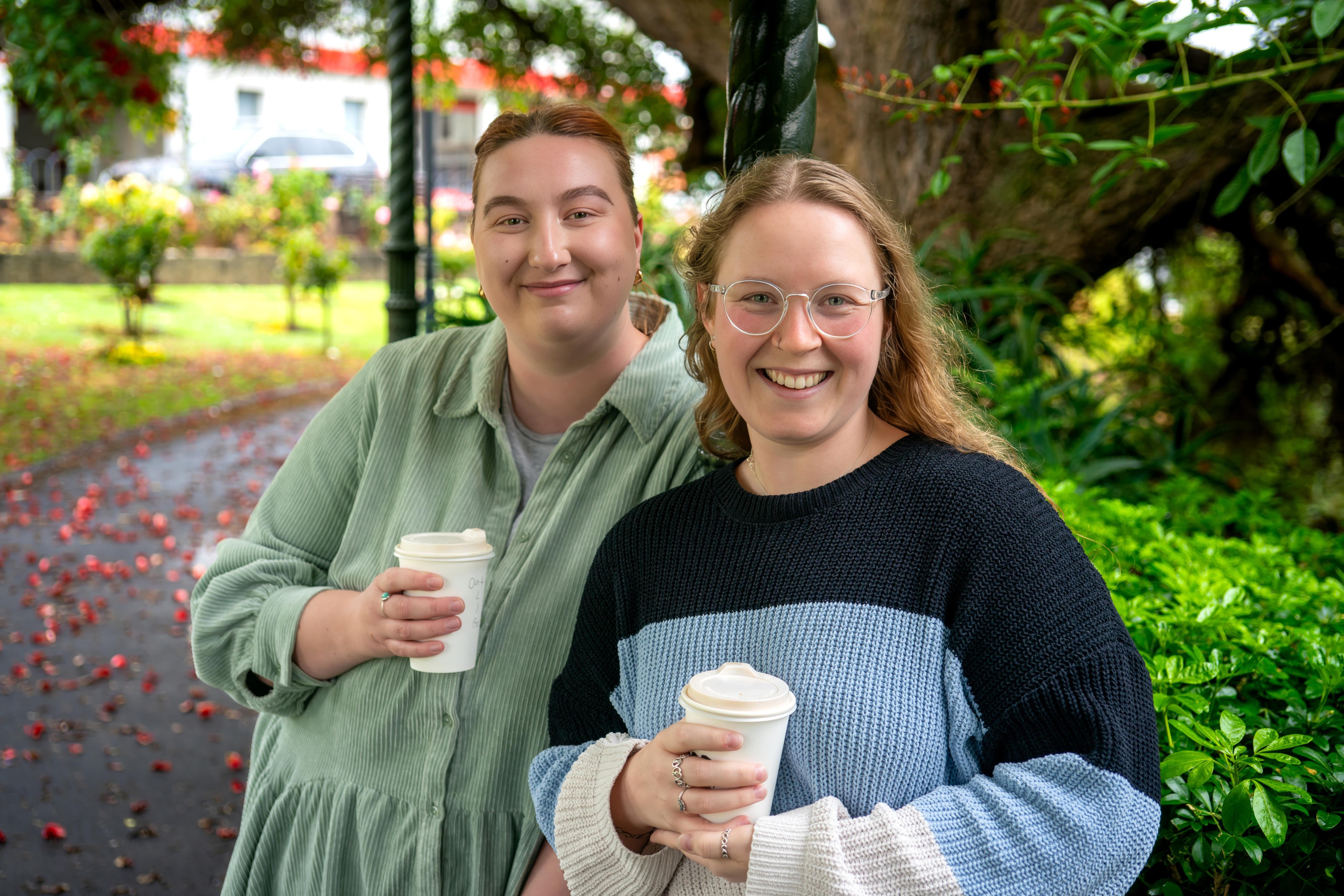 Two women in front of trees smiling with coffee cups